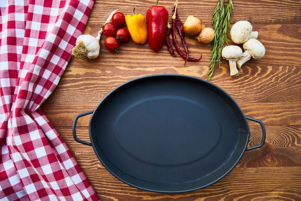 Empty black cast iron pan on a wooden table with tomatoes, peppers, garlic, rosemary and mushrooms
