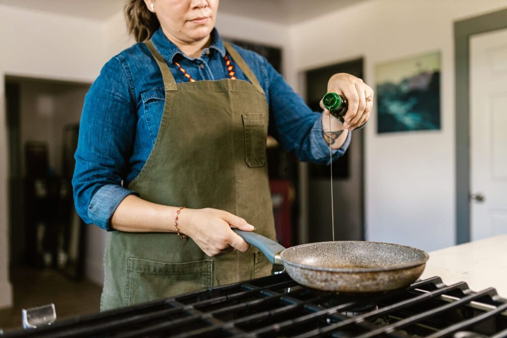 Cook in an apron pouring olive oil from a bottle into a frying pan on a gas stove