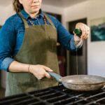 Cook in an apron pouring olive oil from a bottle into a frying pan on a gas stove